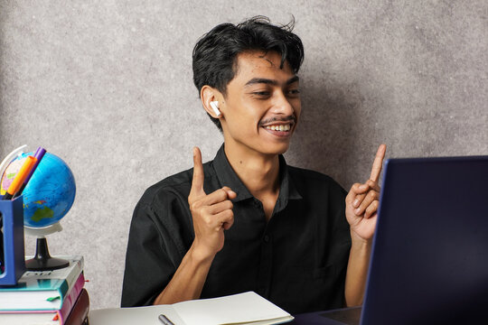 Happy young man in black shirt wearing wireless earphones, smiling and pointing upward with both hands while sitting at a desk with laptop, books, and globe in a modern home office.