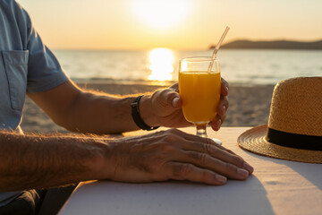 Man relaxing with a glass of orange juice on the beach at sunset. Summer vacation and leisure lifestyle. Close-up of a senior enjoying a holiday.