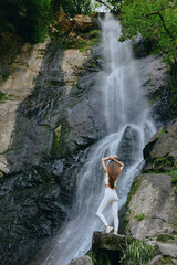 Woman standing near a tall cascading waterfall in white activewear surrounded by lush green forest, showcasing tranquility and natural beauty in an outdoor setting.