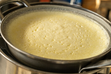 Fresh cow milk heating in a metal pan using boiling water, showing frothy surface and bubbles. Traditional method of preparing natural dairy product in kitchen with warm golden texture.