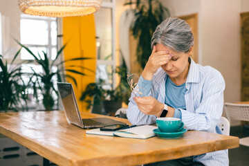 Tired senior woman freelancer holding head in pain while working remotely at cozy cafe