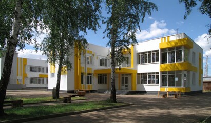 A bright, modern two-story building with white walls and yellow accents, set amidst trees and a grassy area under a blue, partly cloudy sky