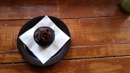 Top view of a chocolate cupcake with dark frosting placed on a napkin inside a black plate over rustic wooden table.  
