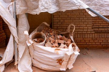 Big bag filled with brick rubble placed under plastic cover against background of brick wall at construction site.