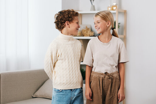 Two smiling children stand indoors in a cozy living room, sharing a friendly conversation. Natural light from windows highlights their cheerful expressions and casual outfits.