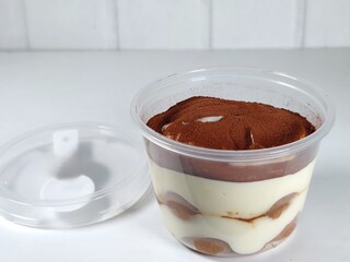 Tiramisu in a plastic cup, the layers are visible, the top has cocoa powder, there is a plastic spoon inside the lid of the plastic cup placed on the side, with a white background.