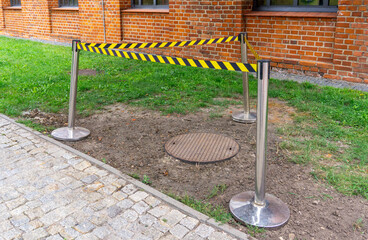 Safety barrier with warning tape securing a manhole cover on a lawn near cobblestone pavement and brick building wall.