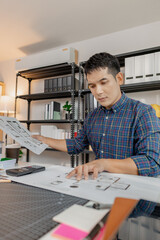Young male architect working thoughtfully at office desk, analyzing house blueprints and construction plans, reviewing details carefully to ensure accuracy in design and successful project planning.