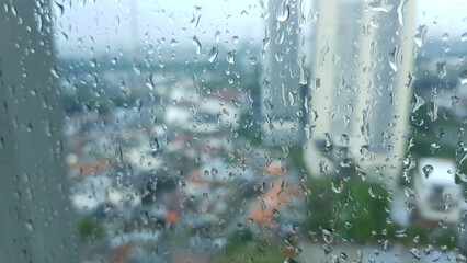 Water droplets on a glass window during rain, with an out-of-focus view of tall buildings and city houses in the background.  
