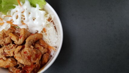 Top view of a rice bowl filled with stir-fried chicken, shredded vegetables, lettuce, and mayonnaise on a dark textured background.  
