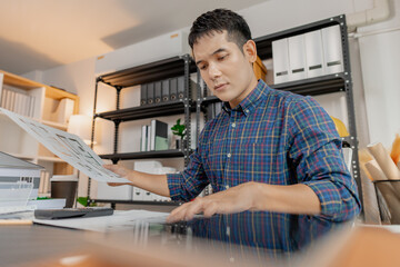 Young male architect working thoughtfully at office desk, analyzing house blueprints and construction plans, reviewing details carefully to ensure accuracy in design and successful project planning.