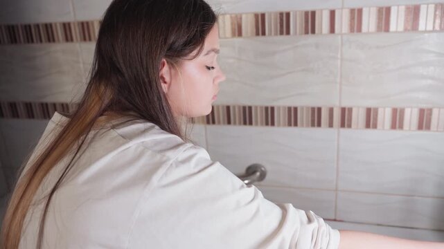 Side view young woman in bathtub reaches chrome mixer, turns off shower tap, arm extended over rippled tiles, soft light on skin, calm hygiene routine, tidy bathroom corner during everyday wash prep