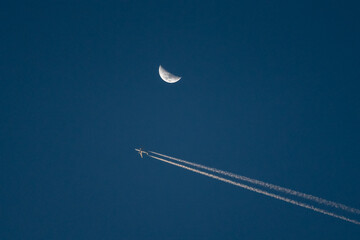 Airplane flying past the moon with contrails in clear blue sky, travel and aviation concept