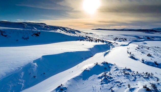 A sunlit aerial view of a vast snow-covered mountainous region, showcasing a winding road and distant lake under a vibrant sky