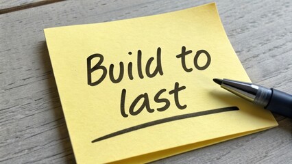 Inspirational yellow sticky note with handwritten text Build to last and black pen on wooden desk background, focus on motivation and goals