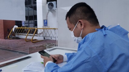 A man wearing a face mask and protective blue gown is sitting at a desk, in a healthcare or testing setting, near a window to the outside.