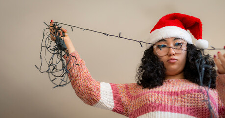 A woman stretching and untangling a bundle of Christmas lights, wearing a Santa hat and a cozy sweater, evoking festive preparation and cheerful holiday spirit. The setting is vibrant and joyful.