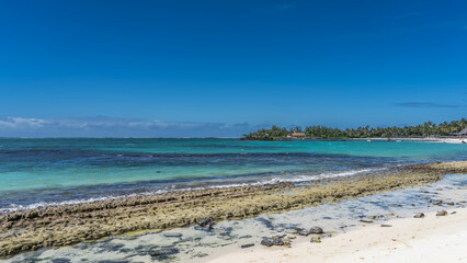 Beautiful seascape. Sandy beach and volcanic rocks by the water. The waves of the turquoise ocean are foaming on the rocks. There are boats, a pier, a hotel building, green vegetation in the distance.