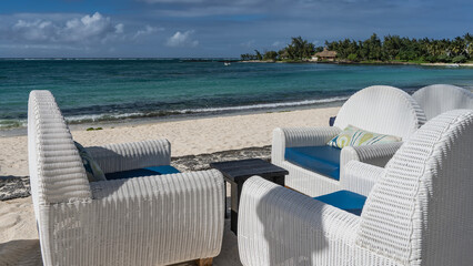 A place to relax. White plastic wicker chairs with cushions on the seat and a coffee table are located on the embankment. The waves of the turquoise ocean are foaming on the sandy beach. Blue sky