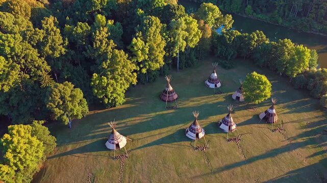Sunset bathes a traditional native american tipi village in golden light, casting long shadows across a grassy clearing amid lush green forest in a dramatic aerial view