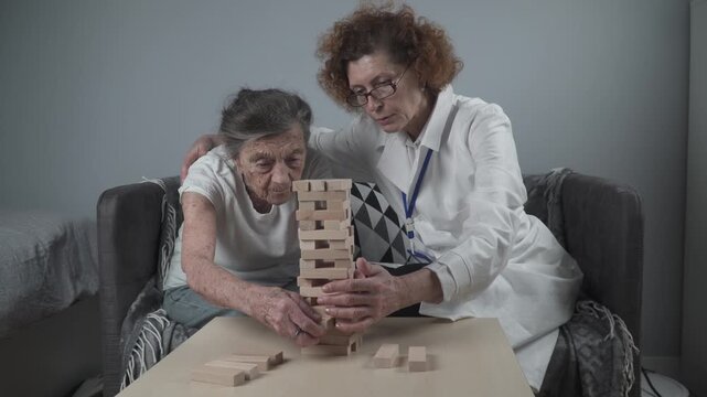 Senior woman practice skills build wooden blocks, building tower and trying not to let it fall, Jenga game. Old patient pull out block, place on top, support dotor during therapy dementia in house.