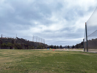 Open golf practice range with green turf, safety nets, scattered golf balls, and hilly landscape on overcast day in Southern California
