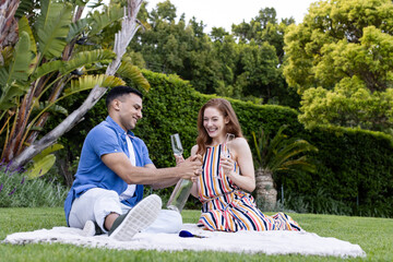 Young couple enjoying picnic outdoors, sharing champagne and smiling on blanket