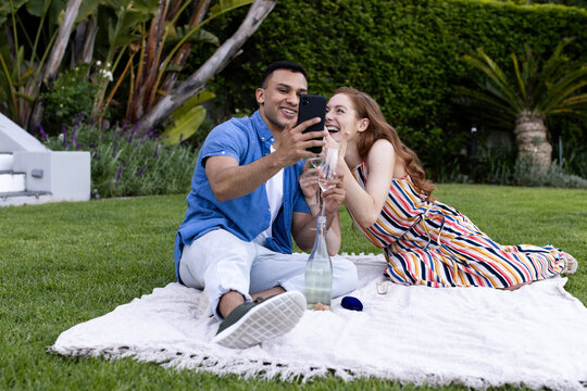Sitting on picnic blanket, couple taking selfie with smartphone, smiling happily - Powered by Adobe