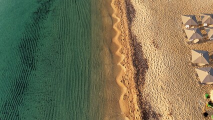 Stunning aerial drone view of a serene beachscape featuring crystal-clear greenish-blue waters gently meeting golden sand. The shoreline forms elegant wave patterns, while neatly arranged white beach 