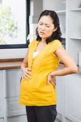 Pregnant Asian woman in kitchen experiencing discomfort, holding back and belly
