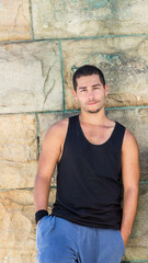 Fototapeta premium Confident man in black tank top leaning against stone wall outdoors