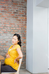 Pregnant Asian woman sitting at home, experiencing back pain, holding belly, copy space