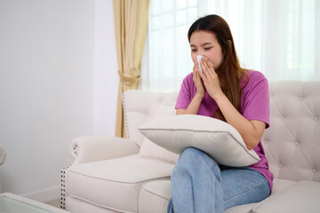 A woman blowing her nose, The scene suggests she's experiencing allergy symptoms, flu, or illness. She is seated on the sofa, feeling unwell