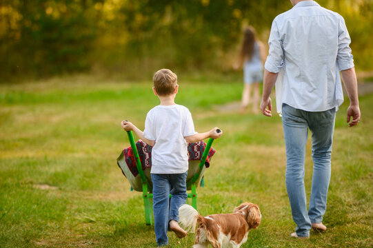 Man walks barefoot through grass, son pushes garden wheelbarrow, and daughter and dog run ahead, getting ready to do some gardening, seen from behind. Gardening concept, family vacation in countryside