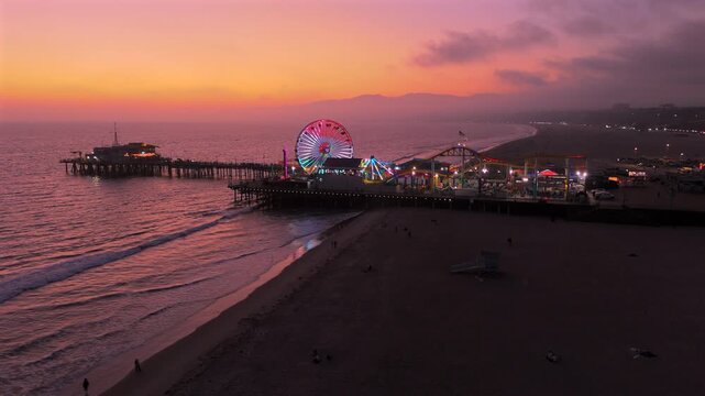 Breathtaking aerial sunset shot showcasing the iconic Santa Monica Pier in Los Angeles, beach, and Ferris wheel, reflecting stunning and vibrant colors as the day turns to night