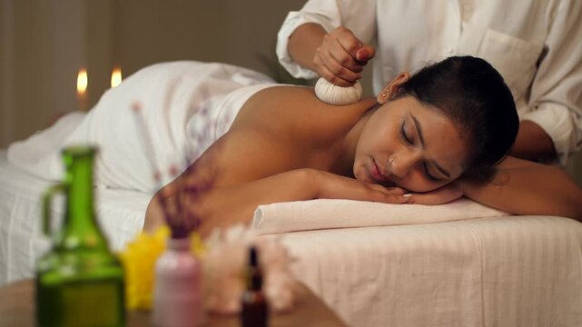 A professional woman masseuse uses herbal compress balls massage on the back of a woman  panchkarma  potli massage. A pan shot of an Indian woman getting Ayurvedic treatment -  treat back pain  mus...
