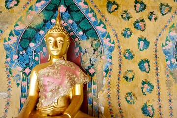 Gold Buddha statue clothed in traditional Thai silk garments with ancient painted backdrop at Wat...