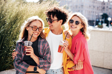 Young three stylish friends posing in the street. Fashion man and two cute female dressed in casual summer clothes. Smiling models having fun. Cheerful women and guy outdoors, Hold and drink lemonade