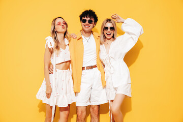 Fototapeta premium Group of young three stylish friends posing in studio. Fashion man and two cute female dressed in casual summer clothes. Smiling models having fun. Cheerful women and guy near yellow wall, sunglasses