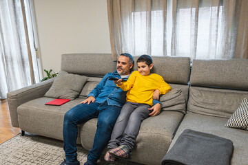 Father and son sitting on a comfortable sofa, watching television and changing channels with a remote control, enjoying time together at home