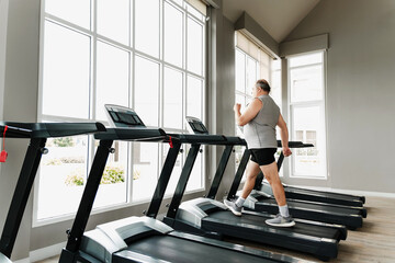 Man walks on treadmill in bright, spacious gym with large windows. Scene shows cardio fitness, wellness, wellbeing, motivation, performance, coaching, strength, running, jogging, and rehabilitation.