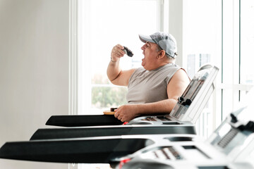 Overweight senior man in casual sportswear sitting on treadmill, eating cake instead of exercising, symbolizing unhealthy lifestyle, wellness challenges, diet, weight loss, and fitness motivation.