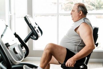 Overweight senior man working out on a recumbent exercise bike indoors, focused on fitness and wellness, symbolizing healthy lifestyle, weight loss, aging health, and active living.