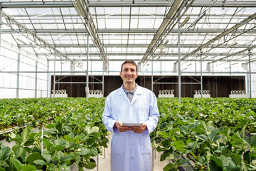 Smiling agronomist in a white lab coat using a tablet while standing among rows of leafy plants inside a bright greenhouse, illustrating modern smart farming and agricultural research.
