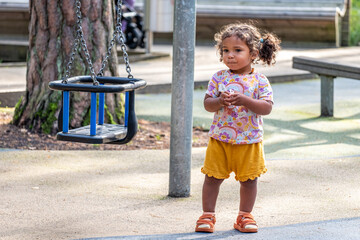 African American girl with curly hair stands playfully beside a swing in a sunny park, embodying...