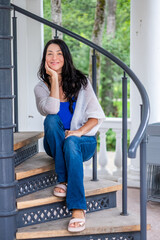 Woman sitting gracefully on staircase, wearing casual attire, smiling joyfully, surrounded by lush...