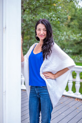 Elegant woman with long dark hair, wearing a blue top and white shawl, poses gracefully outdoors, embodying beauty, relaxation, and a sense of harmony in a serene environment