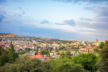 Fototapeta premium Stunning panoramic view of Tbilisi, Georgia, captured at sunset. The city’s colorful rooftops, lush greenery, and rolling hills blend beautifully under a glowing sky of Georgia’s capital town