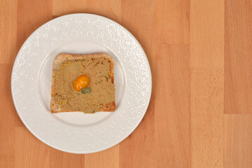 A top-down shot of a piece of toast with homemade chicken liver pate, garnished with a small yellow tomato, on a plate on a wooden table.