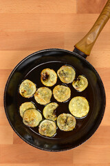 Zucchini slices frying in a pan with oil, placed on a wooden surface